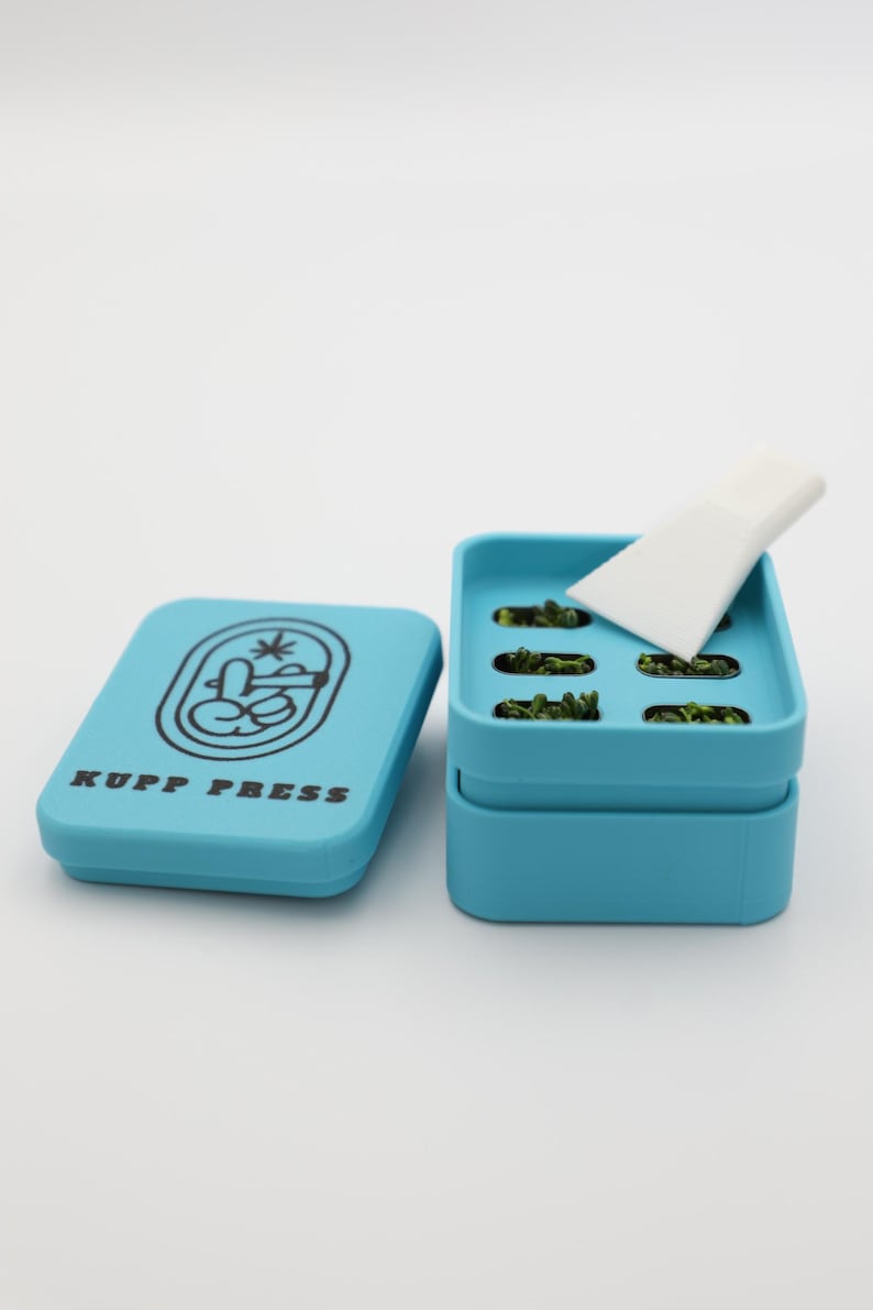 Op de afbeelding: Een lichtblauwe KUPP PRESS-container met een deksel met een logo en tekst. De container is open en onthult verschillende groene items en een wit gereedschap. De container is ontworpen om kleine items in te bewaren.