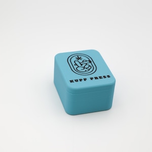 Op de afbeelding: Een lichtblauwe, rechthoekige doos met afgeronde hoeken. De bovenkant van de doos heeft een zwarte afbeelding en de tekst "KUPP PRESS" in zwart. De doos staat op een wit oppervlak.