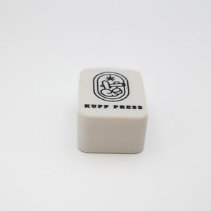 Op de afbeelding: Een klein, rechthoekig, lichtbeige object met afgeronde hoeken. De bovenkant heeft een zwarte afbeelding en de tekst "KUPP PRESS". De afbeelding is een gestileerd ontwerp met een handgebaar en een plant.