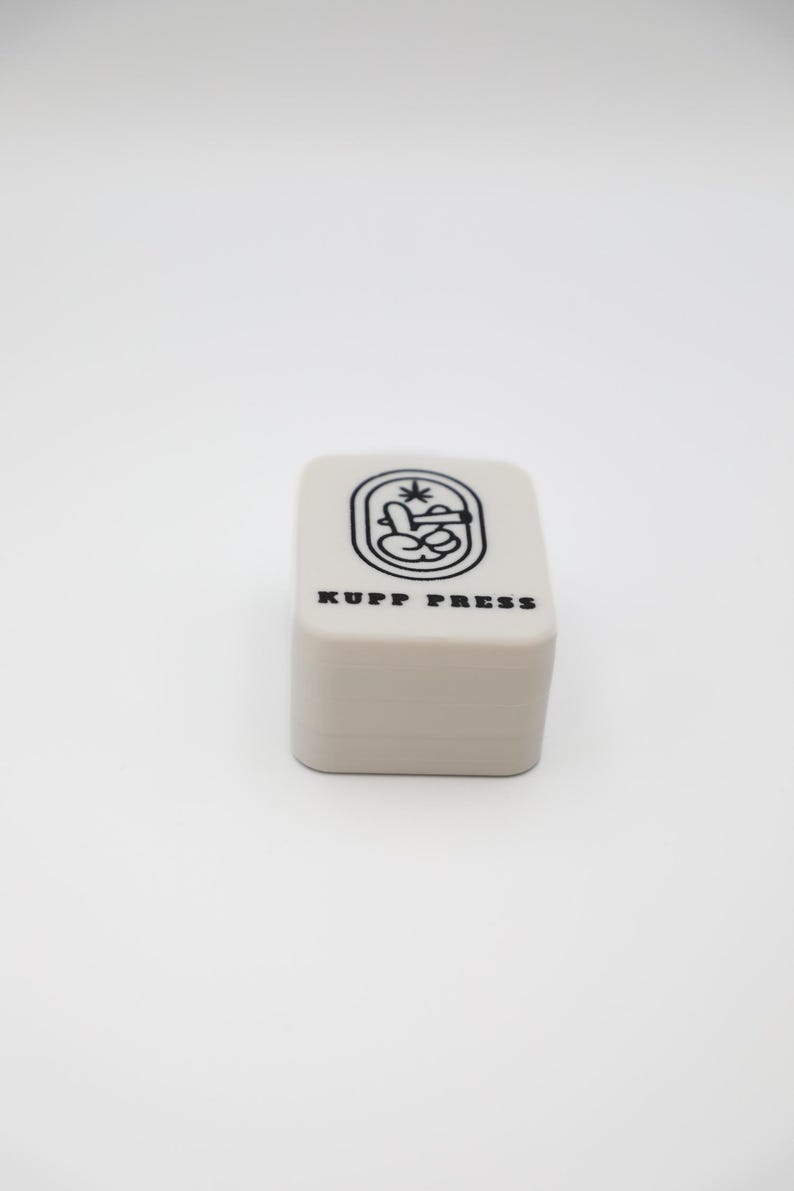 Op de afbeelding: Een klein, rechthoekig, lichtbeige object met afgeronde hoeken. De bovenkant heeft een zwarte afbeelding en de tekst "KUPP PRESS". De afbeelding is een gestileerd ontwerp met een handgebaar en een plant.