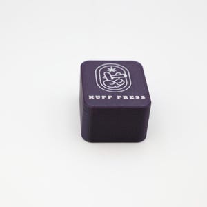 Op de afbeelding: Een donkerpaarse doos met de tekst "KUPP PRESS" in het wit. De doos heeft een witte afbeelding op de bovenkant. De afbeelding heeft een ovale vorm met een ontwerp erin. De doos staat op een wit oppervlak.