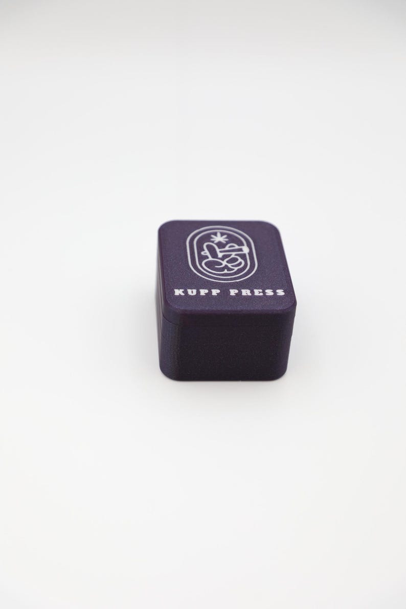 Op de afbeelding: Een donkerpaarse doos met de tekst "KUPP PRESS" in het wit. De doos heeft een witte afbeelding op de bovenkant. De afbeelding heeft een ovale vorm met een ontwerp erin. De doos staat op een wit oppervlak.