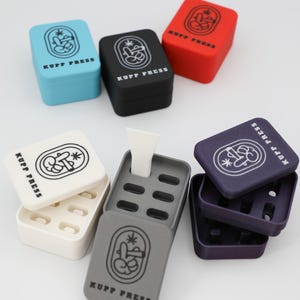 Op de afbeelding: Een assortiment kleurrijke, vierkante KUPP PRESS-containers. De containers zijn in verschillende kleuren, waaronder blauw, zwart, rood, wit, grijs en paars. Elke container heeft de tekst "KUPP PRESS" en een grafisch ontwerp.