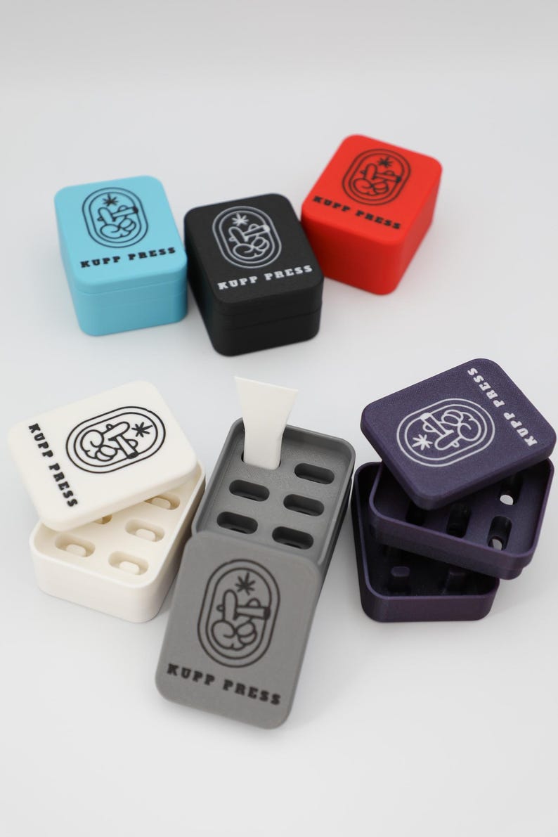Op de afbeelding: Een assortiment kleurrijke, vierkante KUPP PRESS-containers. De containers zijn in verschillende kleuren, waaronder blauw, zwart, rood, wit, grijs en paars. Elke container heeft de tekst "KUPP PRESS" en een grafisch ontwerp.