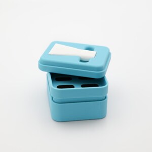 Op de afbeelding: Een lichtblauwe, rechthoekige container met een deksel, gedeeltelijk open. Het deksel heeft een sleuf en een wit, gevouwen papier is zichtbaar. De container is gemaakt van plastic en heeft een eenvoudig, modern ontwerp. De container is waarschijnlijk voor het opbergen van kleine items.