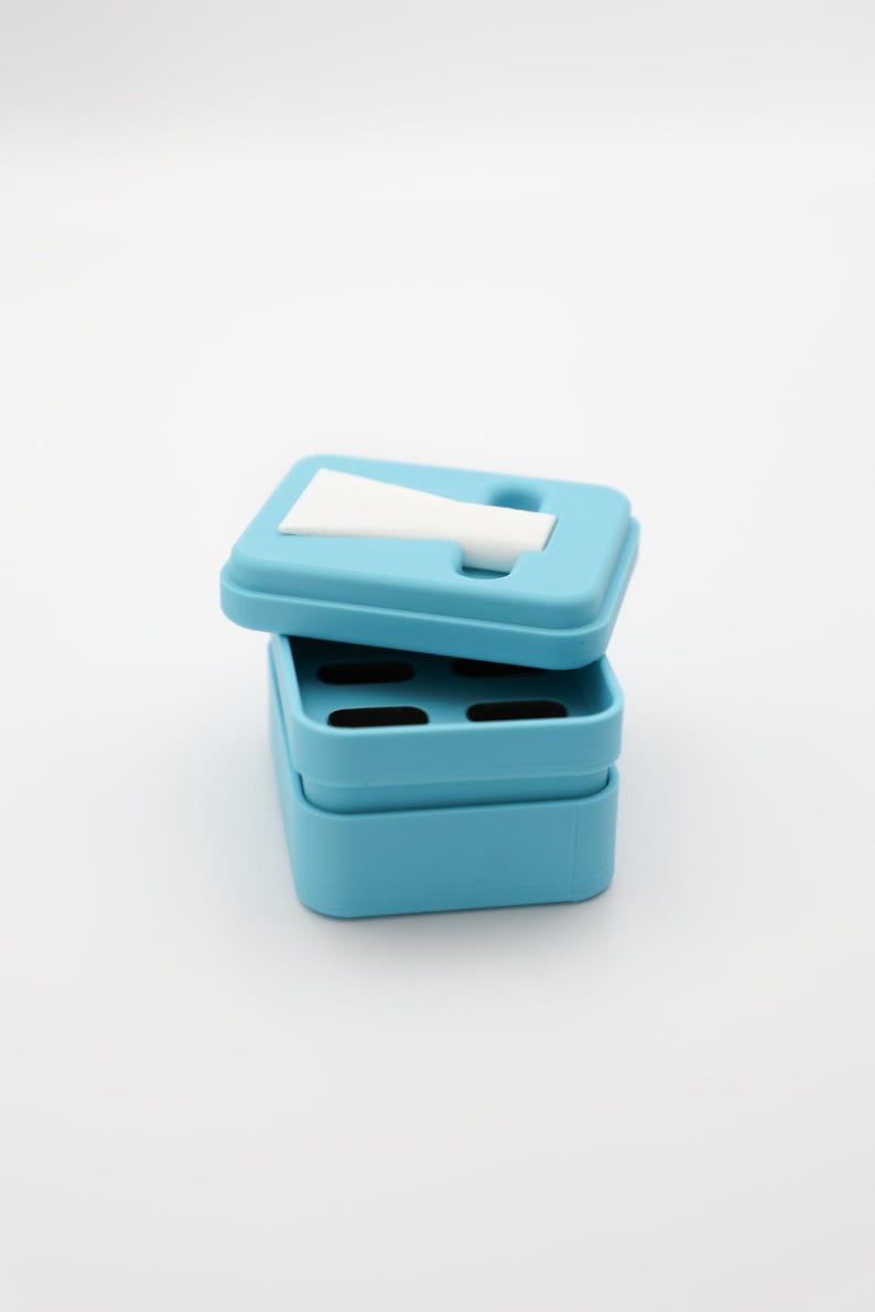 Op de afbeelding: Een lichtblauwe, rechthoekige container met een deksel, gedeeltelijk open. Het deksel heeft een sleuf en een wit, gevouwen papier is zichtbaar. De container is gemaakt van plastic en heeft een eenvoudig, modern ontwerp. De container is waarschijnlijk voor het opbergen van kleine items.