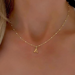 Può includere: Una collana color oro con un piccolo ciondolo a forma di lettera "A". La catena della collana presenta piccole perle distanziate in modo uniforme. Il ciondolo pende dal centro della catena.