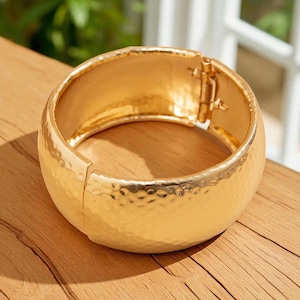 Puede incluir: Un brazalete ancho de color dorado con una textura martillada. El brazalete tiene un cierre de bisagra para facilitar su uso. La superficie refleja la luz, resaltando el diseño texturizado. Una pieza de joyería llamativa.