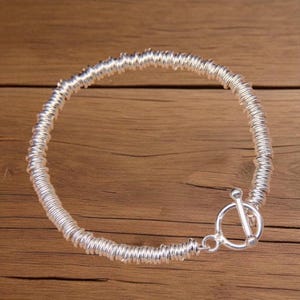 Puede incluir: Una pulsera plateada con un diseño en espiral y un cierre de palanca. La pulsera está hecha de un material metálico brillante y se muestra sobre una superficie de madera. El diseño de la pulsera presenta una serie de espirales enrolladas.