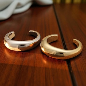May include: Two open-ended, curved metal bracelets. One is silver, the other gold. The bracelets are on a dark wood surface. The bracelets have a smooth, polished finish and a rounded shape.
