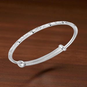 May include: A silver adjustable bangle bracelet. The top of the bracelet has a textured design with star-like shapes. The bottom portion has a series of parallel lines. The bracelet is set against a brown background.