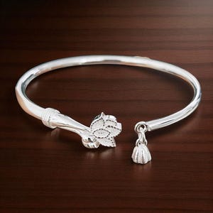 May include: A silver-colored bangle bracelet with a floral design on one end and a small bell-shaped charm on the other. The bracelet is open and rests on a dark brown wooden surface.