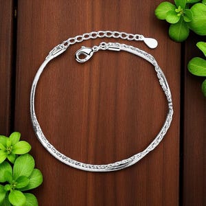 May include: A silver-colored anklet with a snake chain design. The anklet features a lobster clasp closure and an adjustable chain. The jewelry is displayed on a dark brown wooden surface, with green leaves in the background.