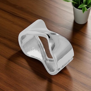 May include: A polished silver bracelet with an unusual, open design. The bracelet has a smooth finish and a curved shape. The item is displayed on a wooden surface.