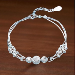 May include: Silver bracelet with a delicate design. The bracelet features multiple strands and decorative beaded accents. The central section includes three textured spheres. The bracelet is displayed against a brown background.