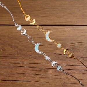 Könnte beinhalten: Zwei filigrane Armbänder mit Mondphasen-Designs. Eines ist silberfarben, das andere goldfarben. Jedes Armband zeigt Halbmonde und kreisförmige Akzente mit schillernden, opalartigen Steinen. Die Armbänder werden auf einer Holzoberfläche präsentiert.