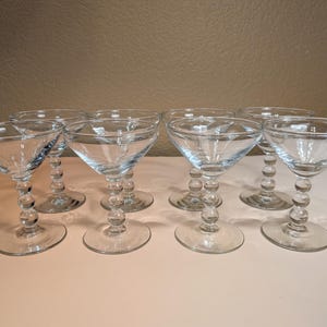 Könnte beinhalten: Acht klare Champagner-Coupe-Gläser mit runden, perlenbesetzten Stielen und breiten, flachen Schalen. Die Gläser sind auf einer weißen Oberfläche angeordnet, die das Licht reflektiert. Das Glaswaren ist ideal zum Servieren von Cocktails oder Sekt.
