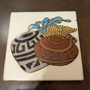 May include: Square ceramic tile featuring a Southwestern design. The tile depicts two pottery vessels in brown, gray, and black, along with corn and blue ribbons. The background is off-white with small brown speckles.