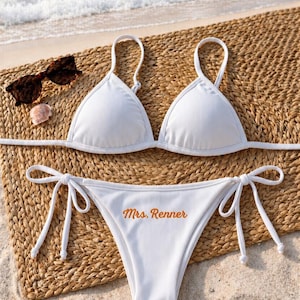 May include: White ribbed bikini set with triangle top and tie-side bottoms. The bottoms feature the text "Mrs. Renner" in orange. Tortoiseshell sunglasses and a seashell are also present.