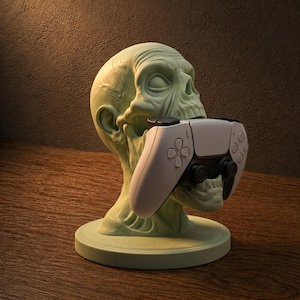 Peut inclure: Un support de manette de jeu en forme de crâne vert pâle. Le crâne tient une manette de jeu blanche et noire dans sa bouche. Le support est sur une base ronde, et le fond est marron foncé.