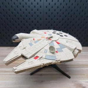 Puede incluir: Un modelo detallado de la nave espacial Millennium Falcon, con un cuerpo color crema y detalles en gris y rojo. El modelo se exhibe sobre un soporte negro, mostrando su diseño icónico y sus intrincados detalles.