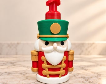 Puede incluir: Un dispensador de jabón rojo, verde y blanco con forma de soldado cascanueces. El dispensador cuenta con una bomba roja, un sombrero verde con un emblema circular dorado, ojos negros, una barba blanca y detalles dorados.