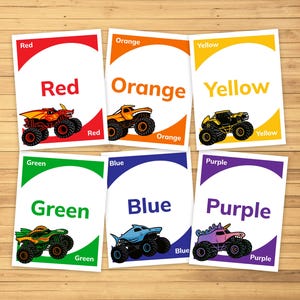May include: Set of seven colorful flashcards with the words "Red", "Orange", "Yellow", "Green", "Blue", and "Purple". Each card features a monster truck in the corresponding color. The cards are on a wooden surface.