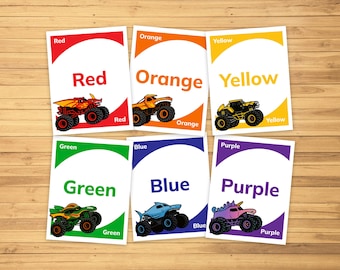 May include: Set of seven colorful flashcards with the words "Red", "Orange", "Yellow", "Green", "Blue", and "Purple". Each card features a monster truck in the corresponding color. The cards are on a wooden surface.