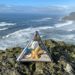 May include: A glass pyramid filled with sand and seashells, including a starfish, on a rocky surface with a view of the ocean in the background.