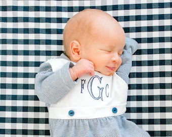 May include: A baby wearing a blue and white striped sleepsuit with a white bib that has the monogram "FGC" embroidered on it. The baby is lying on a black and white checked sheet.