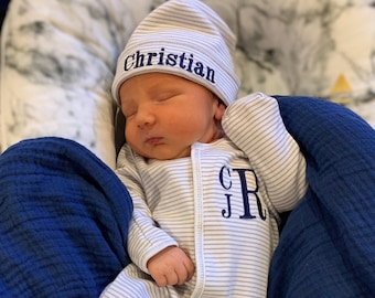 May include: A newborn baby wearing a white and grey striped sleepsuit with the initials "CJR" embroidered on the chest. The baby is wearing a white knitted hat with the name "Christian" embroidered on the front. The baby is lying on a blue blanket.