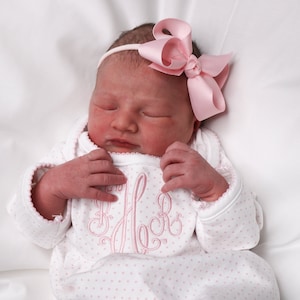 May include: A newborn baby wearing a white onesie with pink polka dots and a pink monogram. The onesie has long sleeves with scalloped edges. The baby is also wearing a pink bow headband. The baby is lying on a white surface.