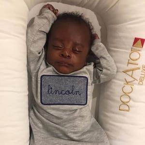 May include: A sleeping baby wearing a gray onesie with a blue embroidered patch that says "lincoln". The baby is lying on a white and gold pillow with the word "docka tot" embroidered on it.