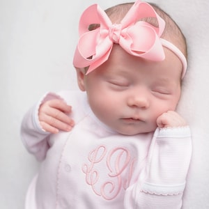 May include: A pink baby onesie with a pink embroidered monogram. The onesie has a white trim around the neckline and sleeves. The baby is wearing a pink bow headband.