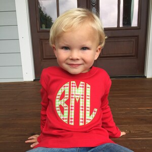 May include: A red long-sleeved shirt with a green, white, and pink plaid monogram in a circle on the front. The monogram reads "BML". The shirt is worn by a young child sitting on a wooden surface.