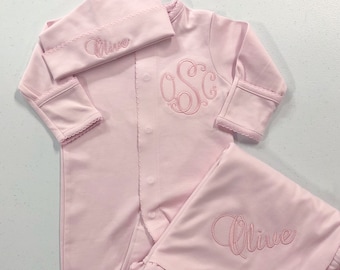 May include: A pink baby outfit set with a hat, onesie, and blanket. The hat and blanket are embroidered with the name "Olive" and the onesie has a monogram "OSC".