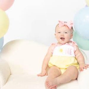 Pode incluir: Um bebé com um macacão amarelo e um babete branco com a inscrição "ONE" está sentado num pequeno sofá branco. Balões em tons pastel, incluindo rosa, azul, verde e amarelo, estão agrupados em torno do bebé, criando um ambiente festivo.