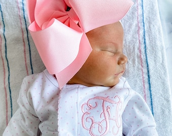 May include: A newborn baby wearing a white onesie with pink polka dots and a pink monogram. The baby is wearing a large pink bow on their head and is sleeping.