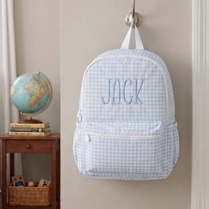 May include: A light blue and white gingham backpack featuring the name JACK embroidered in blue thread on the front. The backpack has a white top handle, white zippers, and a front pocket, hanging from a silver metal wall hook.