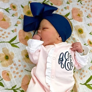 May include: A pink baby onesie with white ruffles and a navy blue monogram. The onesie is worn by a baby with a navy blue bow headband.