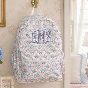 May include: A backpack featuring a repeating pattern of blue flowers and pink bows on a white background. The bag has a front zippered pocket, two side pockets, and white top handles. The letters AWS are embroidered in blue and pink on the front.