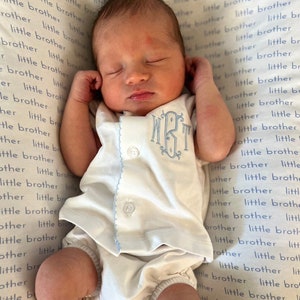 May include: A newborn baby wearing a white romper with light blue trim and monogram. The baby is lying on a blanket with the words "little brother" printed all over it.