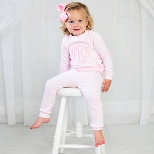 Puede incluir: Un ni&ntilde;o peque&ntilde;o con pijama de rayas rosa y blancas con un monograma rosa en la parte delantera. El ni&ntilde;o est&aacute; sentado en un taburete blanco.