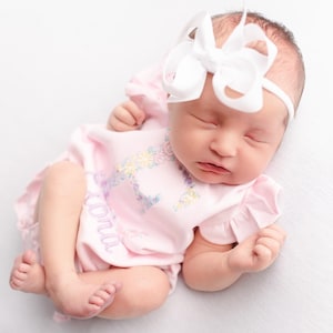 May include: A newborn baby wearing a pink onesie with floral embroidery and the name "Loni" embroidered on the front. The baby is also wearing a white headband with a large bow. The baby is lying down with eyes closed.