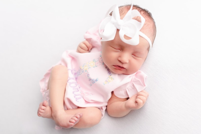 May include: A newborn baby wearing a pink onesie with floral embroidery and the name "Loni" embroidered on the front. The baby is also wearing a white headband with a large bow. The baby is lying down with eyes closed.