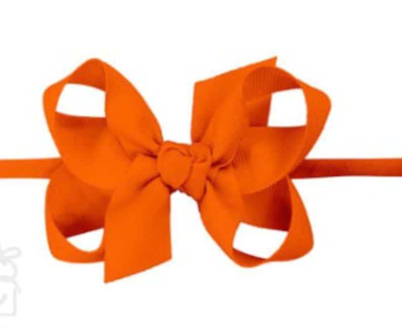 May include: Orange fabric headband with a large bow. The bow is made of a soft, textured fabric and is tied in a simple knot.
