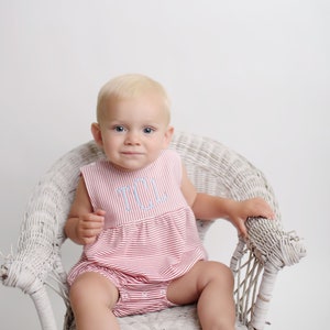 May include: A baby wearing a red and white striped romper with a light blue monogram sits in a white wicker chair. The romper has a sleeveless design and a ruffled hem. The baby has blonde hair and is looking at the camera.