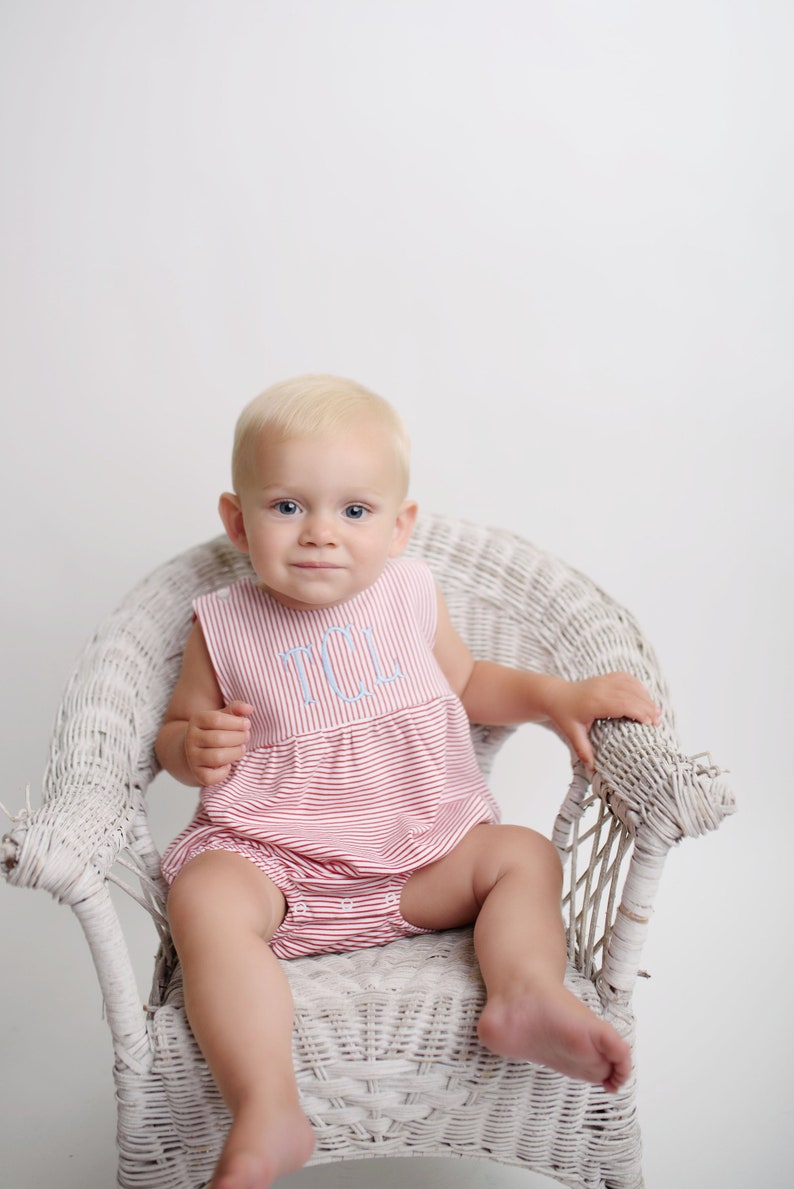 May include: A baby wearing a red and white striped romper with a light blue monogram sits in a white wicker chair. The romper has a sleeveless design and a ruffled hem. The baby has blonde hair and is looking at the camera.