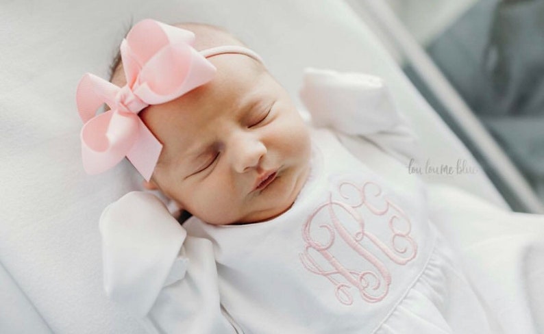 May include: A newborn baby girl wearing a white gown with a pink embroidered monogram and a pink bow headband. The baby is sleeping peacefully.