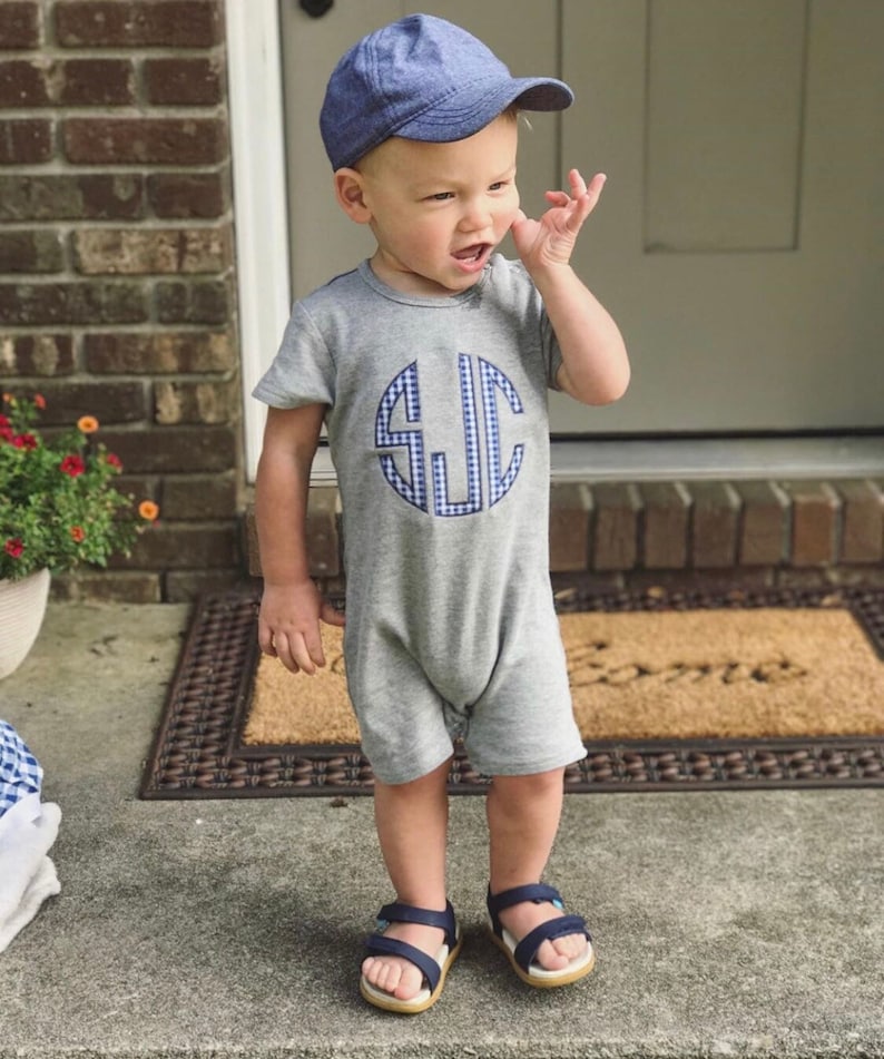 Pu&ograve; includere: Un bambino piccolo che indossa una tutina grigia a maniche corte con un monogramma gingham blu e bianco sul davanti. Il bambino indossa anche un berretto da baseball blu in denim e sandali blu.